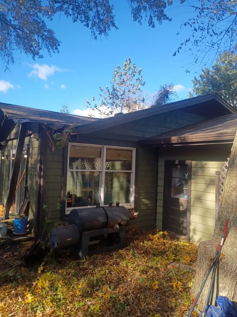 A green house with a dark roof is partially covered in fallen leaves near a large tree trunk